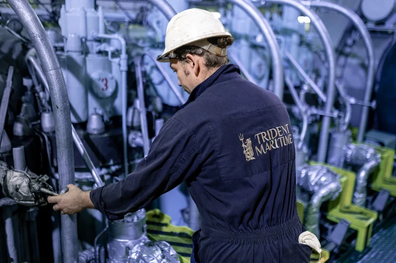 A male crew member in a hard hat and dark blue coveralls inspects large industrial machinery and pipes in an engine room.