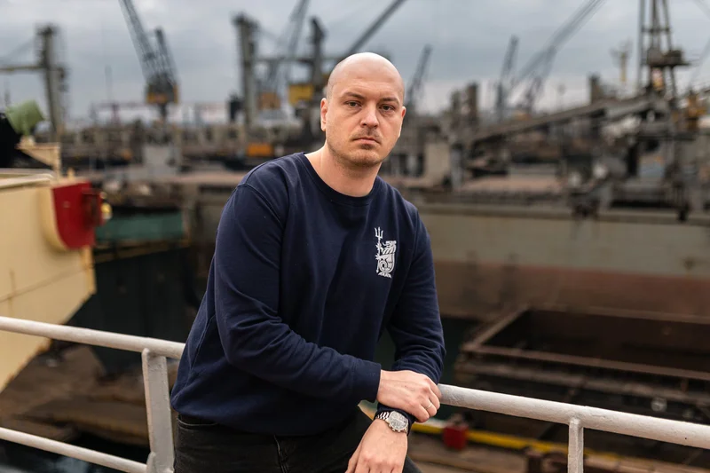 A bald man in a navy sweatshirt leans on a white railing, with multiple ships and port cranes visible in the industrial backg