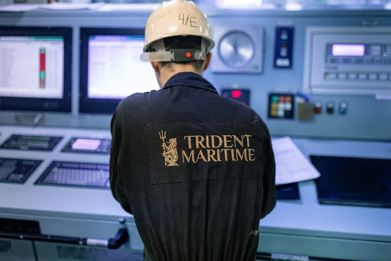 A crew member in a hard hat and dark overall views data on multiple computer screens within a vessel's control room.