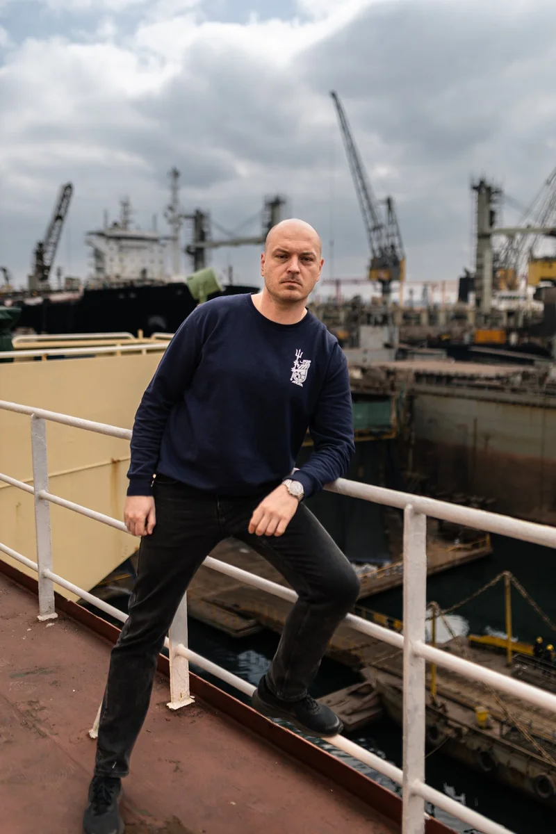 A bald man in a navy sweatshirt and black jeans leans on a railing, with multiple ships, port cranes, and a dry dock visible