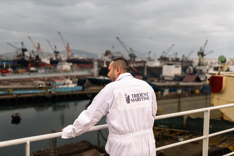 A man in a white jumpsuit stands on a vessel deck, looking towards a busy shipyard with multiple ships and cranes.