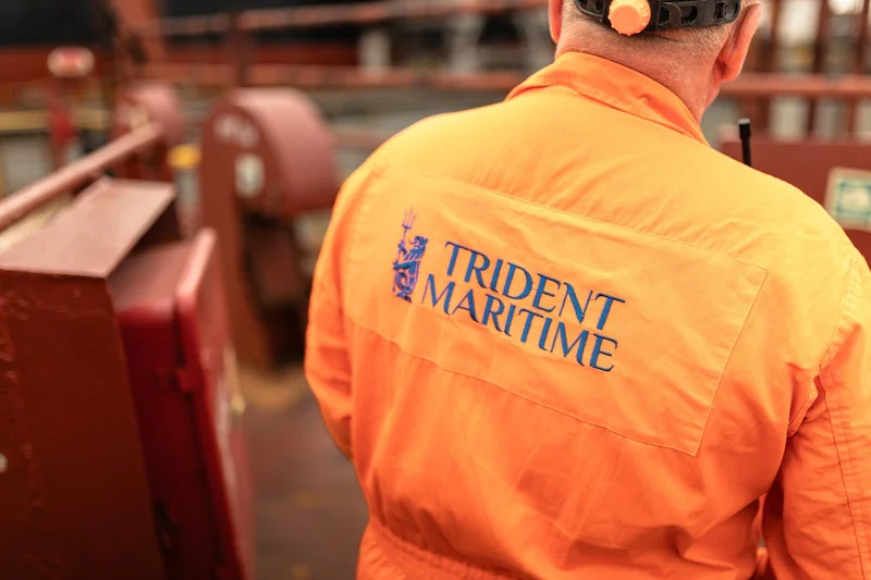 A person's back wearing an orange coverall with the Trident Maritime logo, a hard hat, and blurred red industrial structures