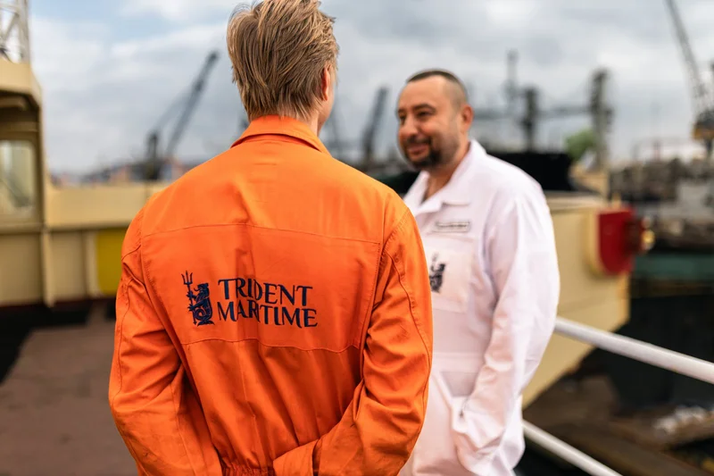 Two men in work jumpsuits on a vessel deck, with industrial port cranes and buildings visible in the blurry background.