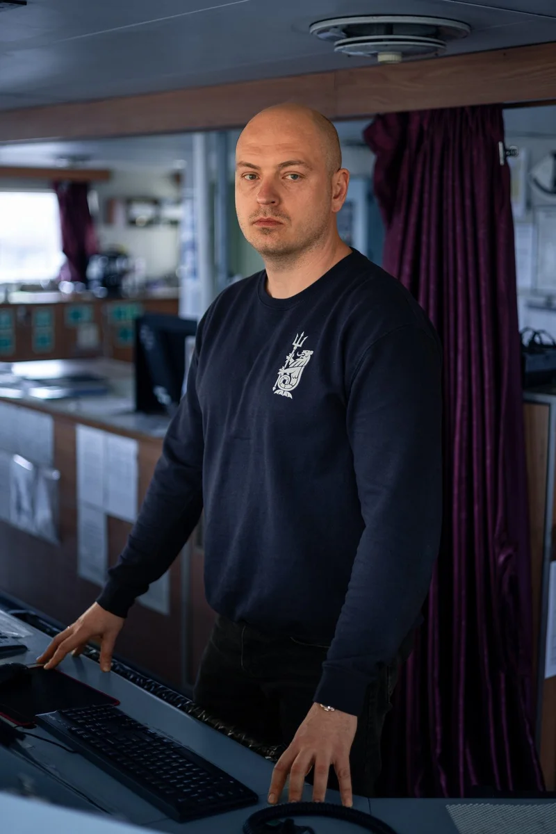 A bald male crew member in a navy blue long-sleeve shirt stands at a control panel with a keyboard on a ship's bridge.