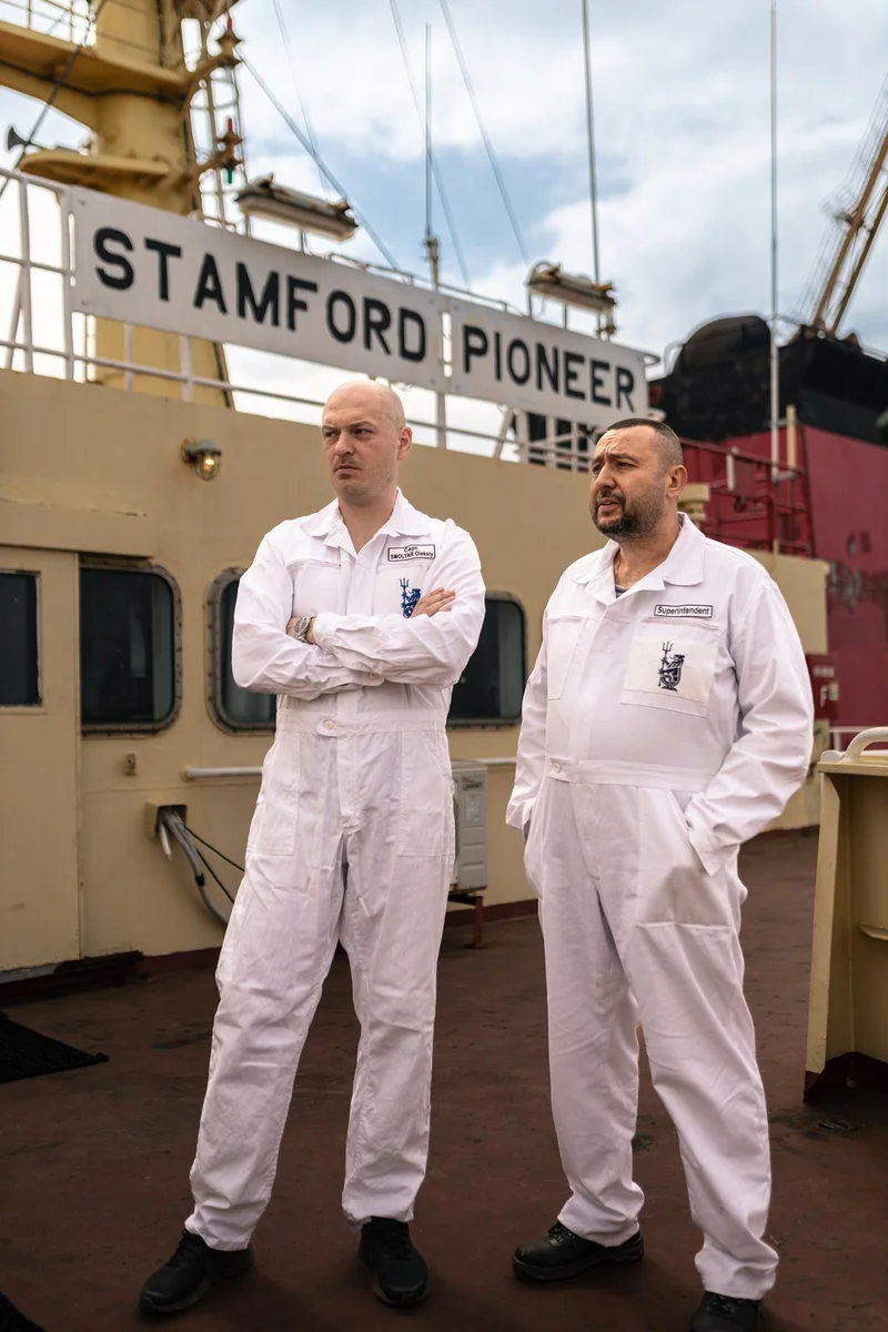 Two male crew members in white coveralls, identified as a Captain and Superintendent, stand on the deck of the vessel "STAMFO