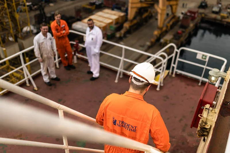 Four men in work overalls and hard hats are on a vessel deck, overlooking a shipyard with cranes and docks.