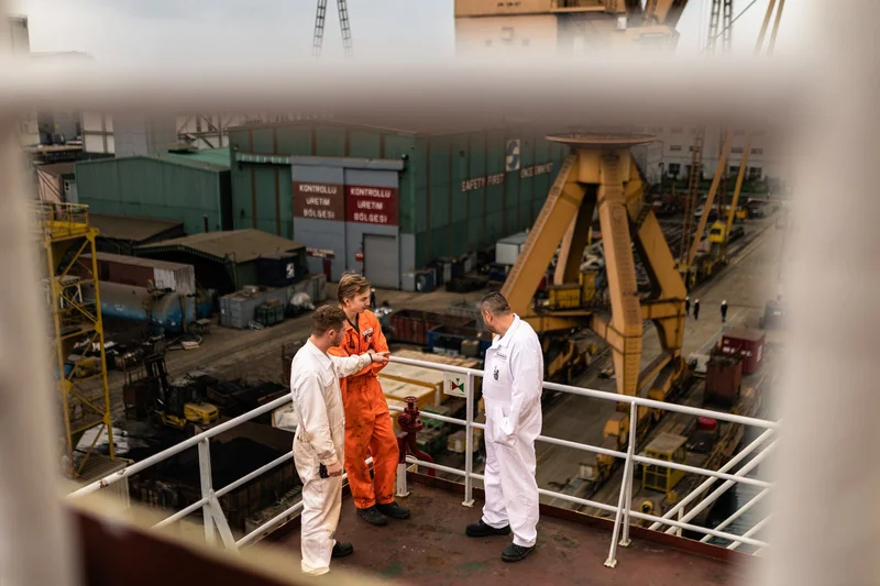 Three crew members, two in white and one in orange overalls, stand on a ship's deck, overlooking a shipyard with cranes and b