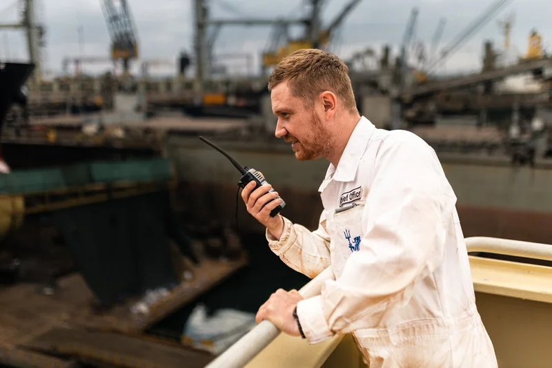 A man in a white overall with a "Chief Officer" patch is holding a walkie-talkie on a vessel railing, with ships and cranes i