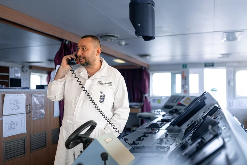 A bearded man in a white 'Superintendent' overall talks on a corded phone on a ship's bridge with control panels and windows.