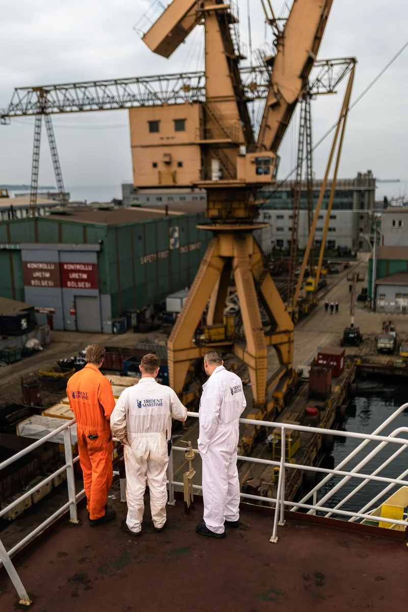 Three men in work jumpsuits stand on a ship deck, overlooking a shipyard with large cranes, industrial buildings, and a dock
