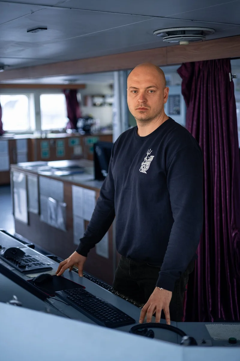 A bald male crew member stands at a ship's bridge control console with keyboards and controls, inside a vessel.
