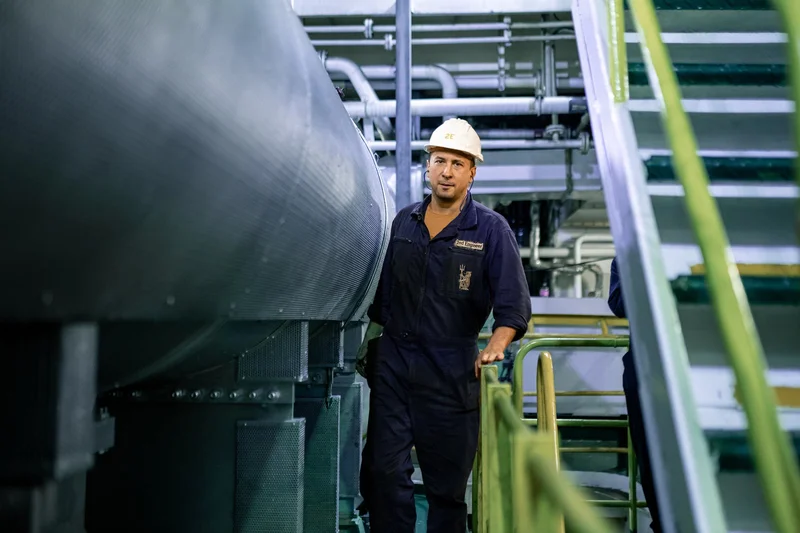 A male 2nd engineer wearing a white hard hat and blue jumpsuit stands next to large machinery and pipes in a ship's engine ro