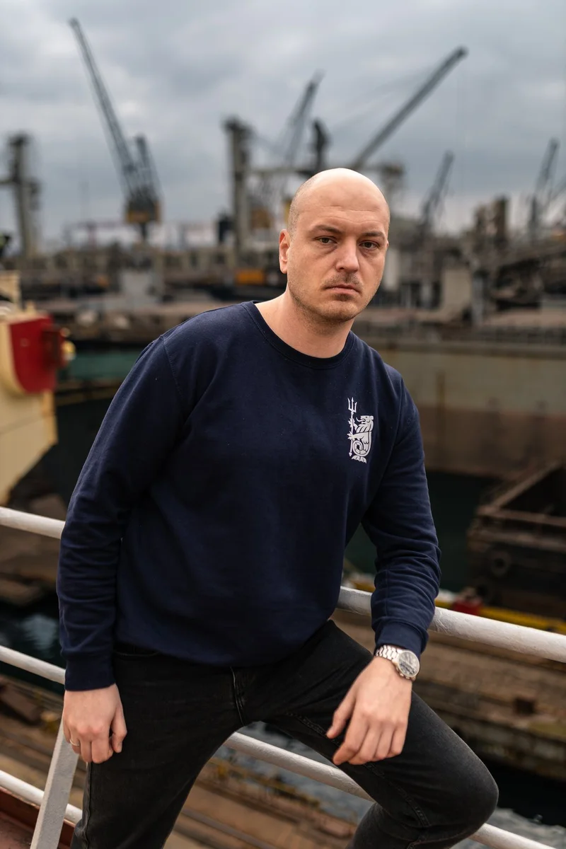 A bald man in a navy blue sweatshirt and jeans leans on a railing with industrial cranes and vessels in a shipyard background