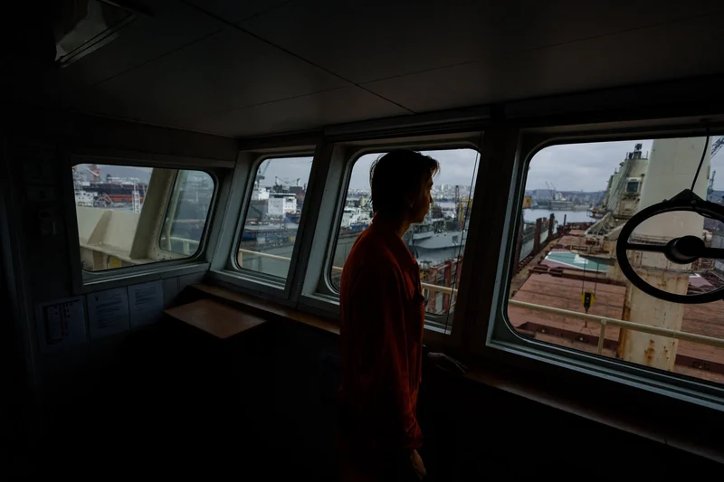 A crew member in a red shirt stands silhouetted on a ship's bridge, looking out at a port with multiple vessels and cranes.