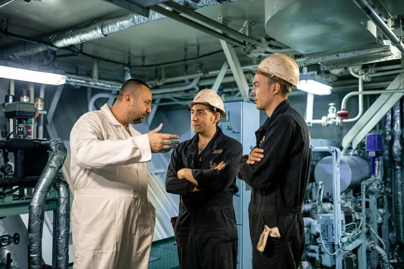 Three male crew members, two in hard hats, discussing in an engine room or industrial space filled with machinery and pipes.
