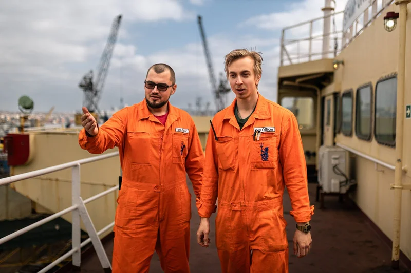 Two maritime officers wearing orange work suits stand on a ship's deck, with port cranes and a city visible in the background