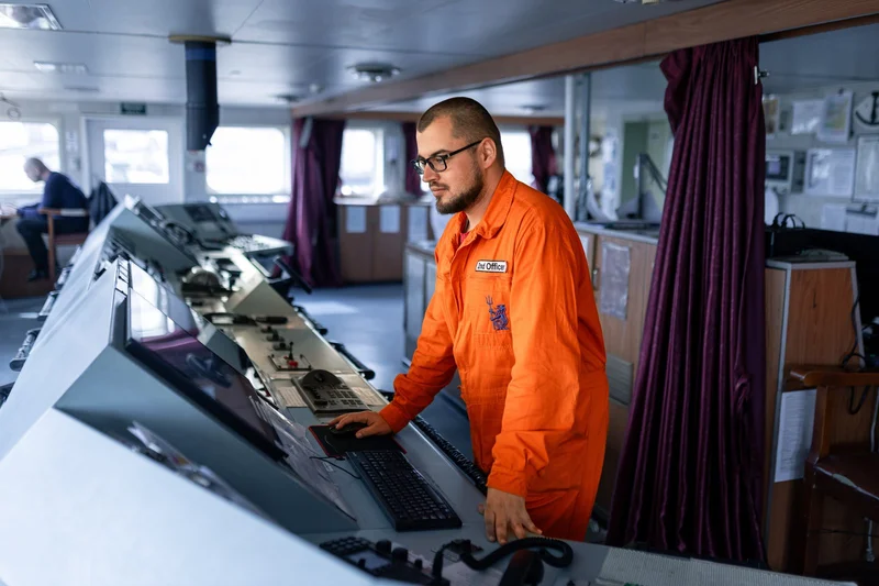 A male crew member wearing an orange uniform is operating a computer and controls on a ship's bridge.