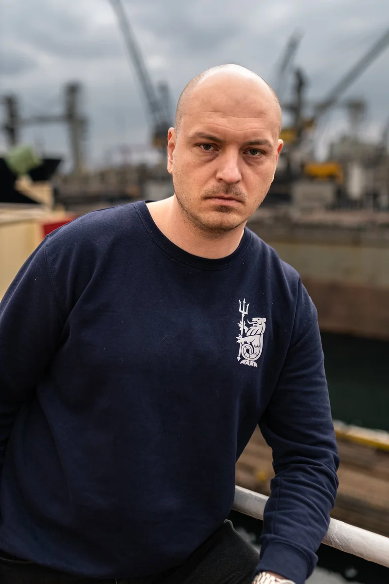 A bald man in a navy blue sweatshirt with a trident emblem stands in front of blurry shipyard cranes and docked vessels.