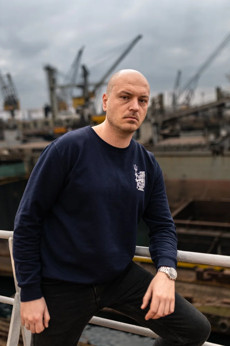 A bald man in a dark blue sweatshirt and black jeans leans on a railing, with industrial cranes and vessels blurred in the ba