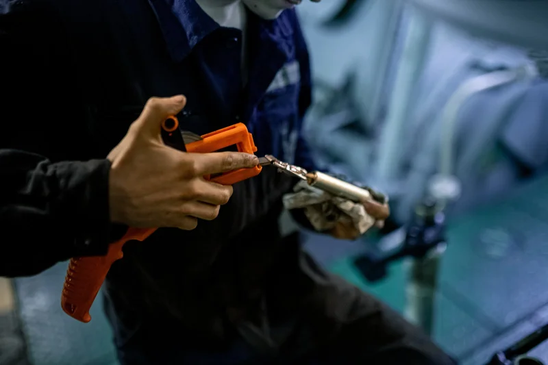 A person's hands use an orange crimping tool on a small metal terminal wrapped in a rag, inside a vessel's interior.