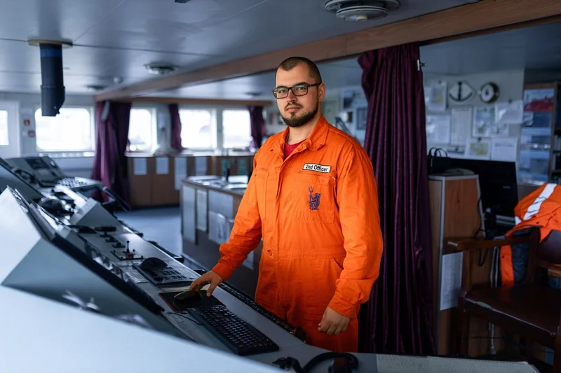 A male 2nd Officer in an orange uniform operates a computer mouse at a ship's bridge control panel with navigational equipmen