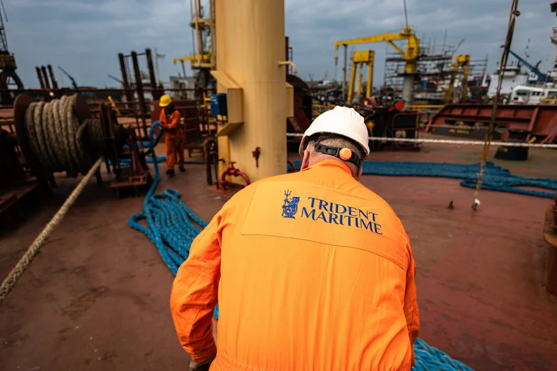 Two crew members in orange jumpsuits and hard hats work on a vessel's deck, managing ropes and winches with a shipyard visibl