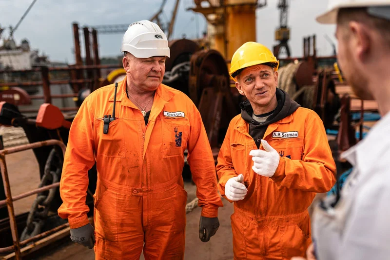 A Bosun and an A.B. Seaman in orange overalls and hard hats converse on a ship deck, with industrial equipment in the backgro