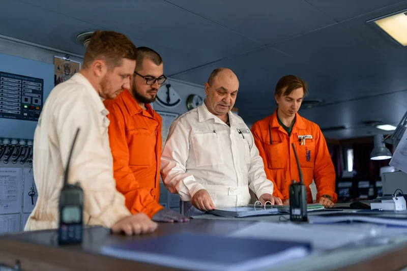 Four male crew members examine documents in a ship's control room, with a two-way radio on the table.