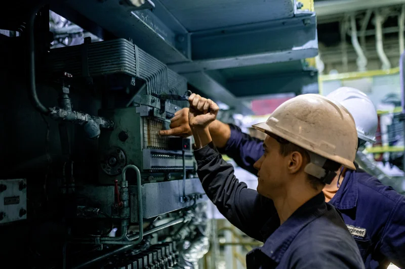 Two engineers in hard hats and work overalls inspecting a large engine or industrial machine in an engine room, one pointing 