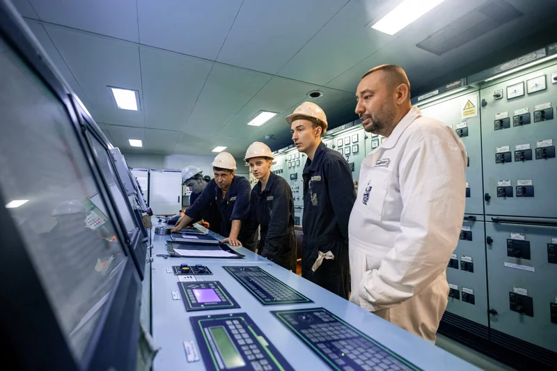 Five crew members, four in hard hats, are in a ship's control room, observing a large control panel and an electrical switchb