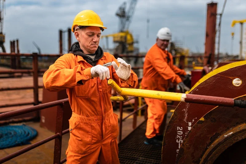 Two crew members in orange coveralls and hard hats work on a ship deck, one adjusting gloves near industrial equipment.