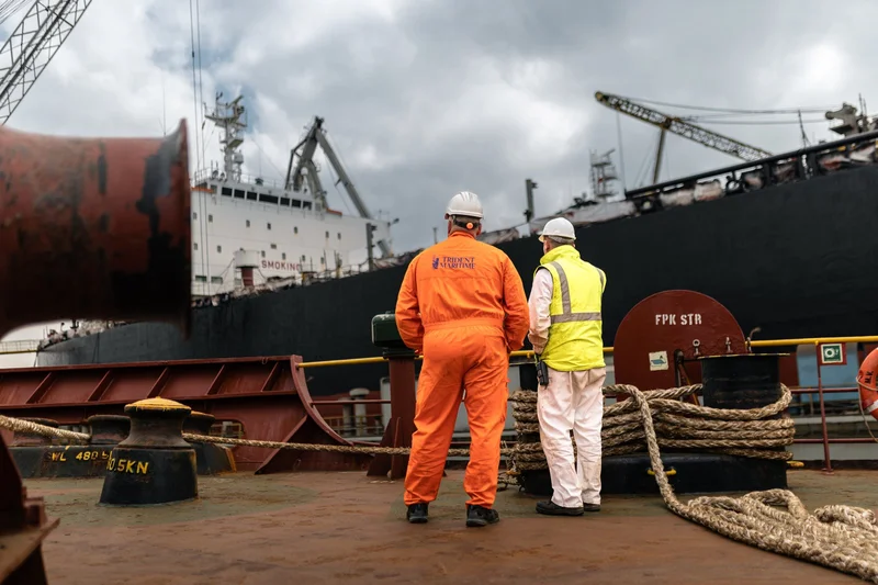 Two workers in hard hats and safety gear stand on a vessel's deck, observing a large cargo ship with cranes and coiled ropes.