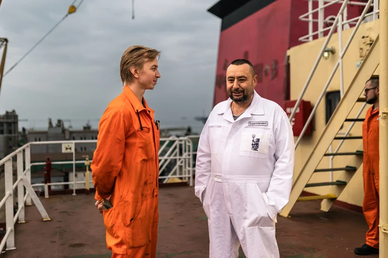 Two men in work overalls stand on a ship deck; one wears orange, another in white has a 'Superintendent' patch. A third man i