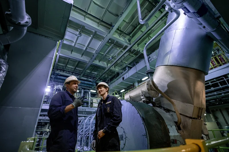 Two crew members in hard hats and overalls talk in an engine room filled with large machinery, pipes, and overhead structures