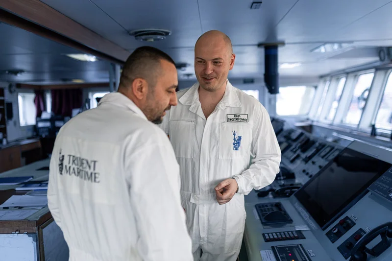 Two male crew members in white uniforms conversing on a ship's bridge, with navigation equipment and large windows visible in