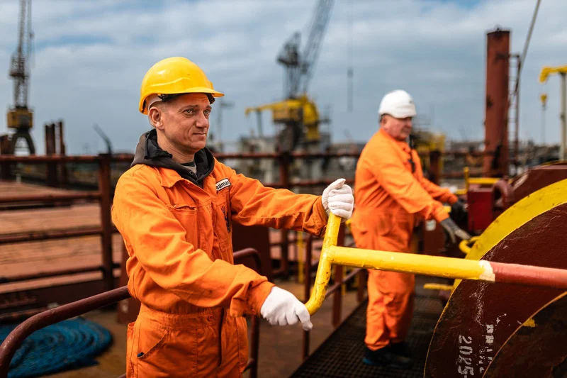 Two crew members in orange coveralls and hard hats operate winches on a vessel deck, with port cranes in the background.