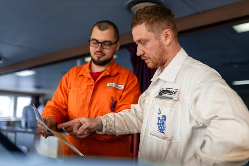 Two male crew members, one a 2nd Officer, wearing coveralls, are reviewing and discussing documents indoors on a vessel.