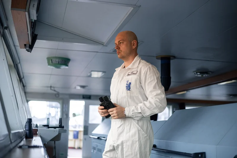 A bald male crew member in a white uniform holding binoculars stands in the wheelhouse of a vessel, looking out the windows.