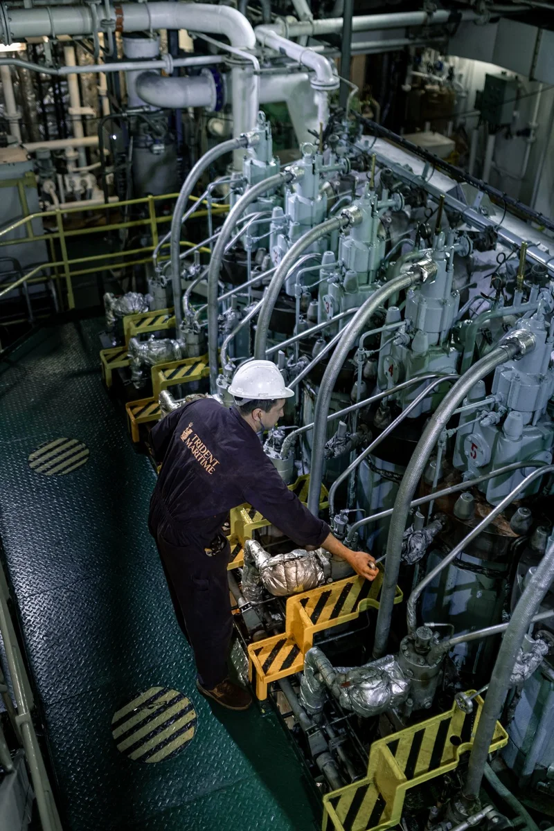 A crew member in a hard hat and uniform inspects a large multi-cylinder marine engine with visible pipes and yellow safety ba