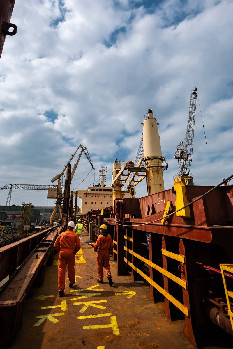 Three workers in hard hats and safety overalls walk on the rusted deck of a large cargo vessel, with ship cranes and port cra