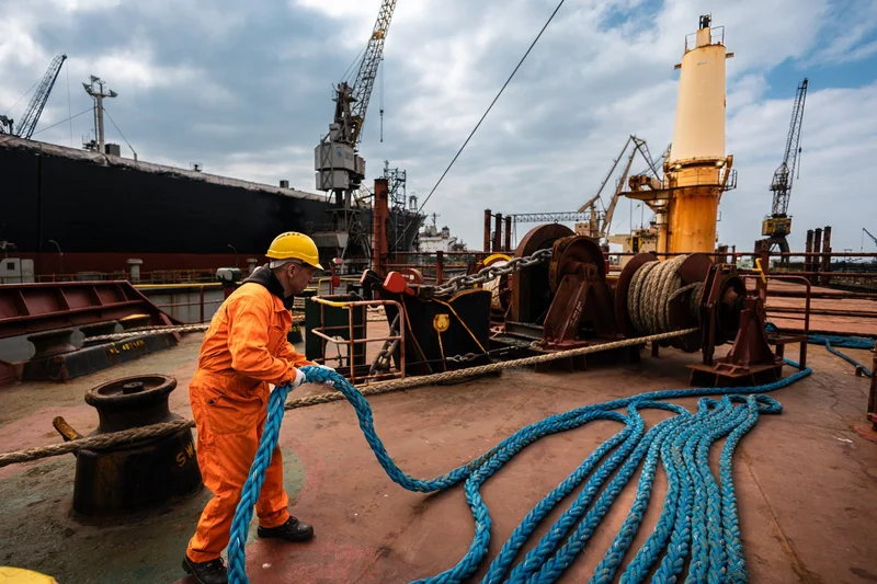 A worker in a hard hat and orange jumpsuit pulls a blue rope on a ship's deck with mooring equipment and shipyard cranes.