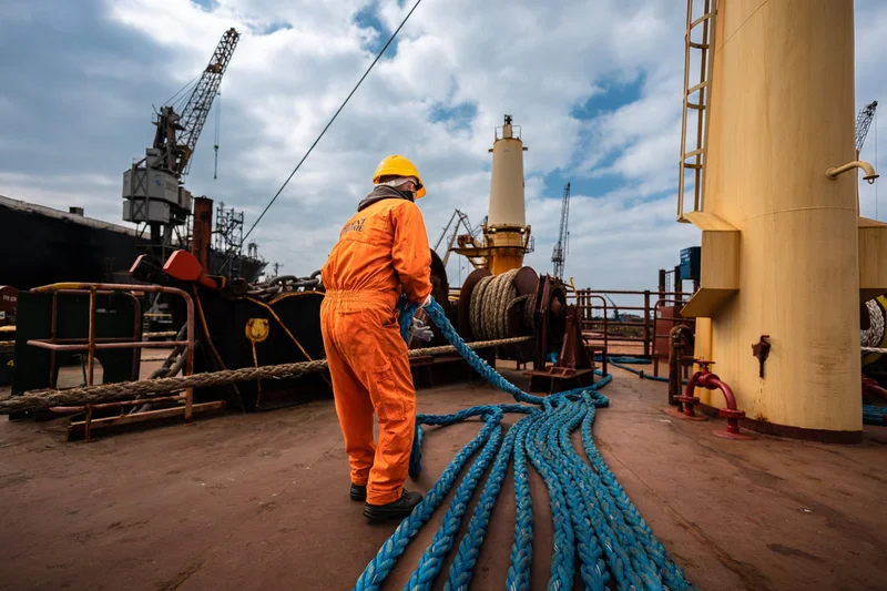 A crew member in orange overalls and a hard hat pulls a blue mooring rope on a ship's deck, with winches and port cranes in t