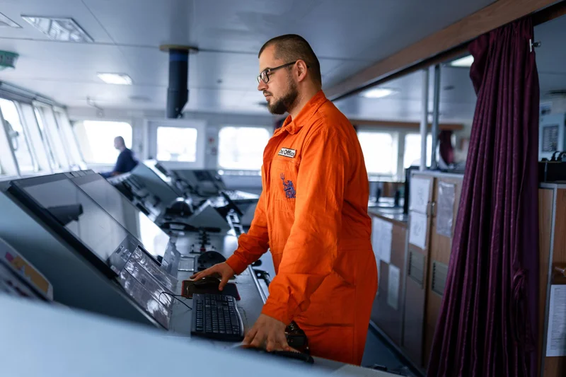 A man in an orange jumpsuit labeled 'Chief Officer' operates controls on a vessel's bridge, with another crew member in the b