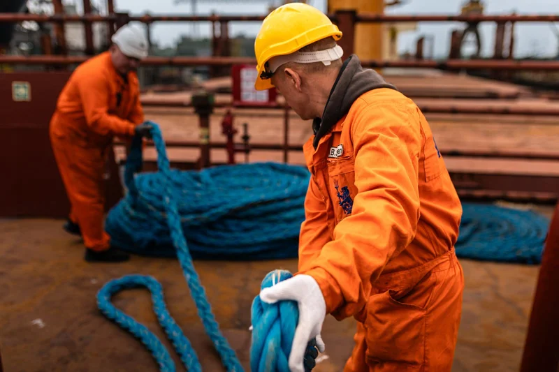 Two crew members in orange overalls and hard hats are coiling large blue ropes on a rusty ship deck during an overcast day.