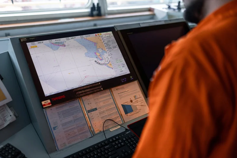 A crew member in an orange uniform stands at a ship's bridge console, viewing a digital navigation chart displayed on a monit