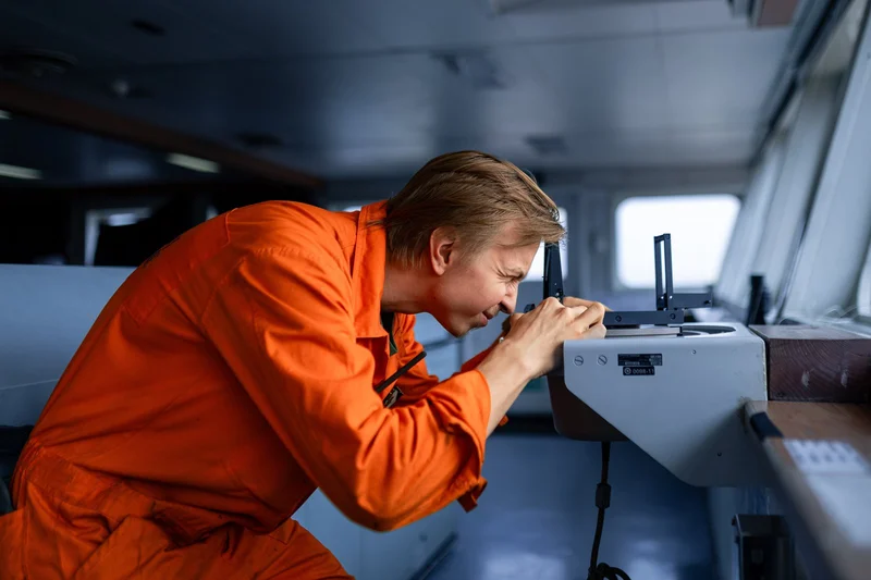 A male crew member in an orange uniform looks intently through a navigation device on a ship's bridge.