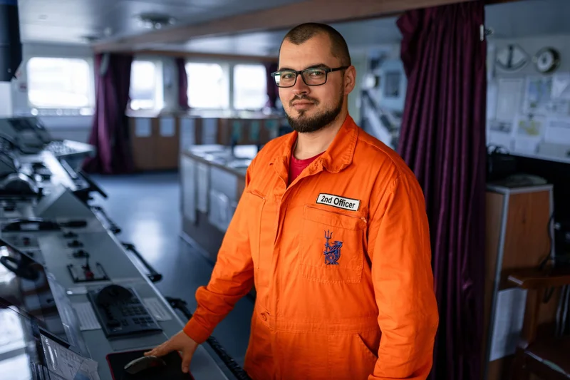 A male 2nd Officer wearing an orange uniform stands on a ship's bridge, with his hand on a computer mouse, surrounded by cont