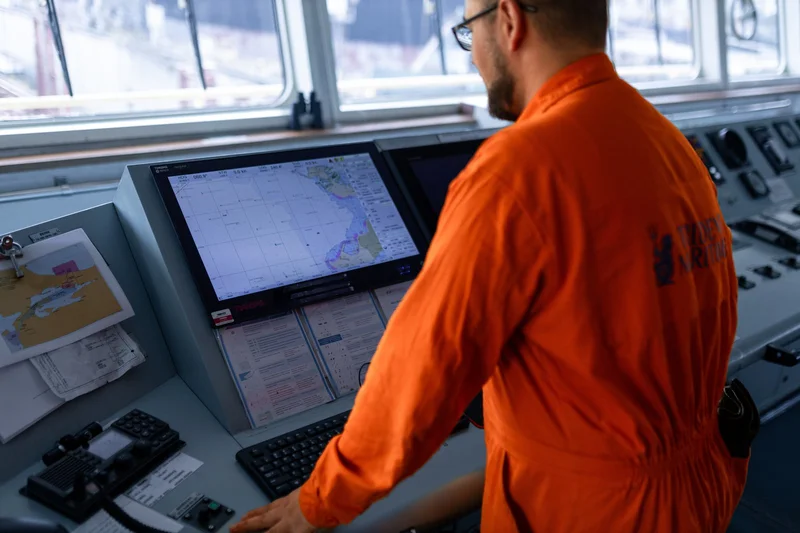 A crew member in an orange jumpsuit stands at a ship's bridge console, viewing a nautical chart on a navigation screen.