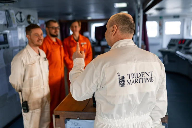 Four crew members in jumpsuits are in a vessel's control room, with one facing and speaking to the other three.