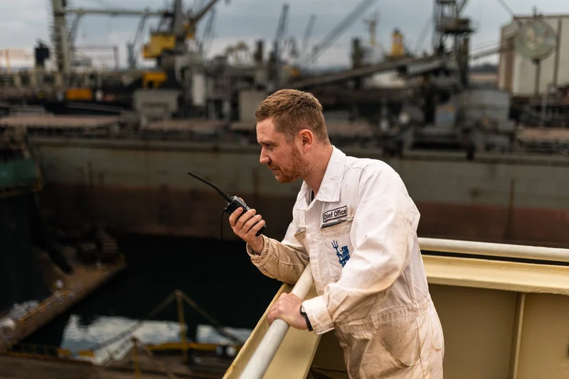 A man in white overalls with a 'Chief Officer' patch holding a walkie-talkie on a ship deck overlooking a dry dock with vesse